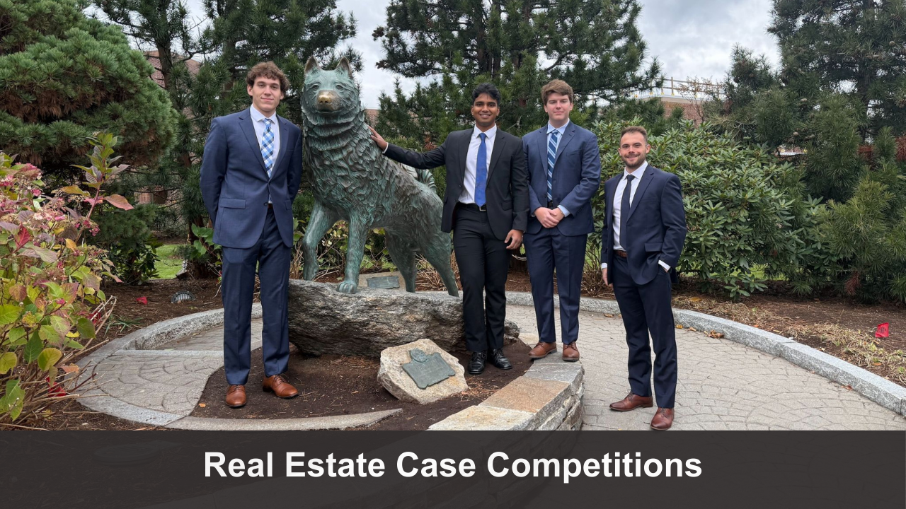 Group of students dressed in business attire standing together next to a dog statue and smiling in an outdoor setting. Banner text reads "Real Estate Case Competitions."